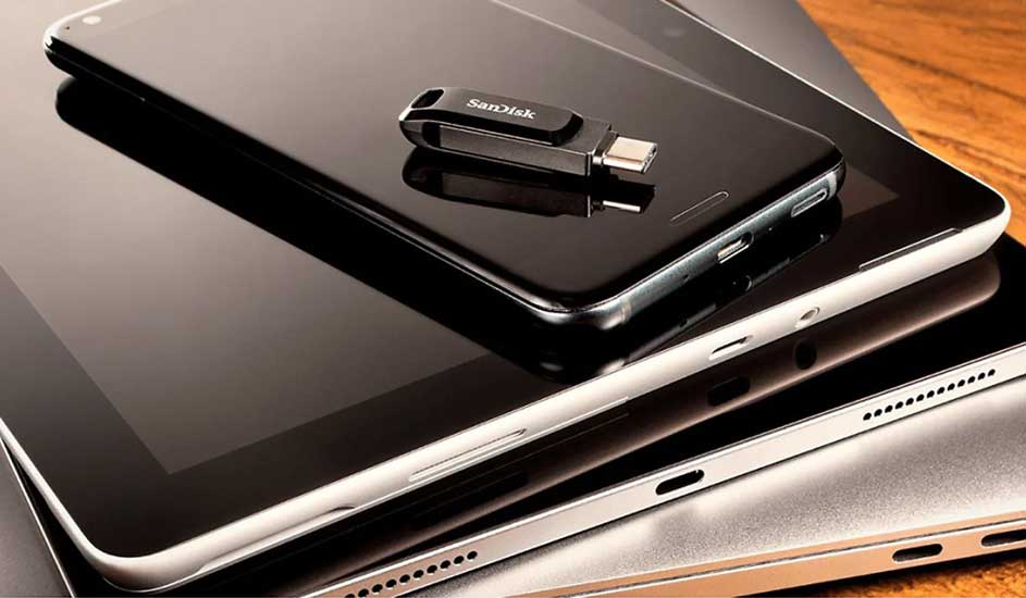 Op een houten tafel liggen een zilverkleurige laptop, een tablet en een zwarte smartphone met een SanDisk USB-stick erop.