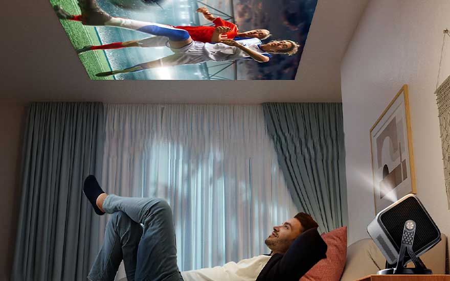 Een man ligt ontspannen op bed terwijl een projector een voetbalwedstrijd groot op het plafond van een gezellige kamer toont.