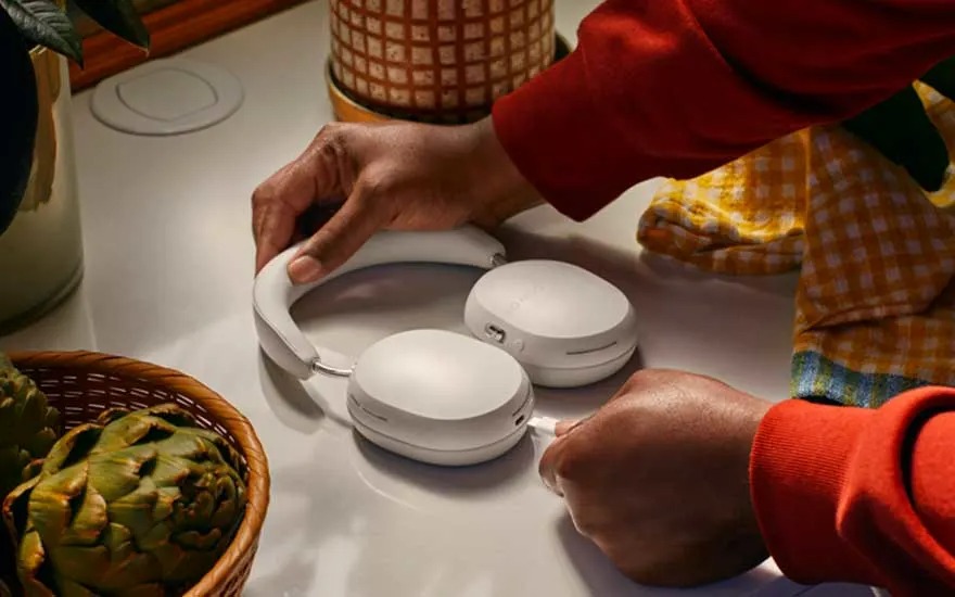 Sur une table, un casque blanc est manipulé pour brancher un câble. Un panier d'artichauts et un pot de plante sont visibles.