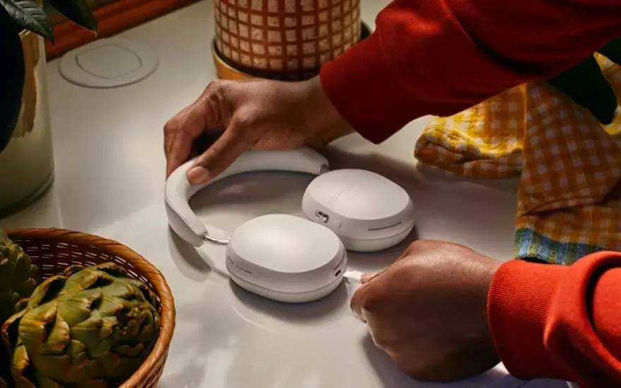 Sur une table, un casque blanc est manipulé pour brancher un câble. Un panier d'artichauts et un pot de plante sont visibles.