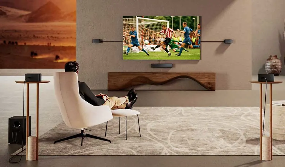 In een ruime, stijlvolle kamer kijkt iemand ontspannen naar een tv met een voetbalwedstrijd, omringd door luidsprekers.