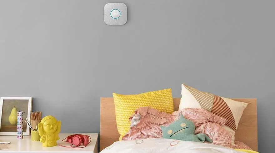Een grijze muur met een witte slimme sensor. Er staat een bed met kussens en een tafel met een foto en decoraties.