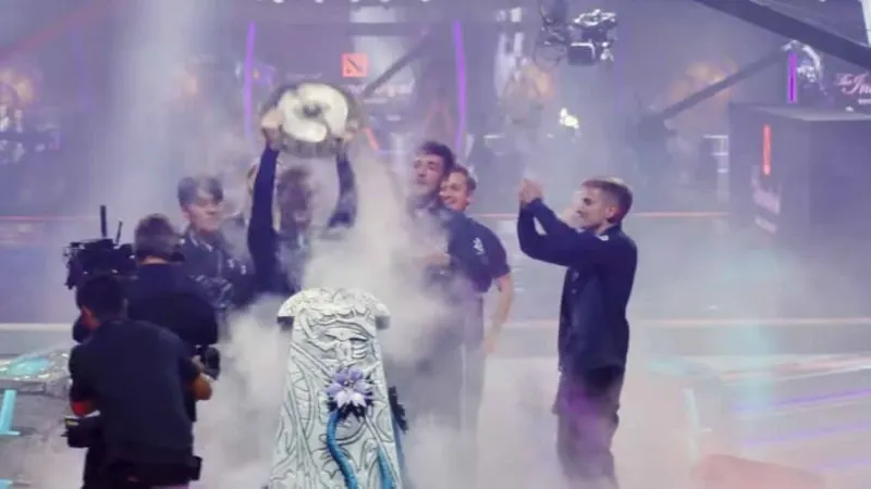 Smoke-filled arena with a trophy being held. Stage with lights and camera present. Foreground features a decorated object.