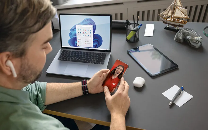 Een bureau met een laptop, tablet, telefoon, pennen en een decoratief zeilschip. Het scherm van de laptop toont een blauwe achtergrond.