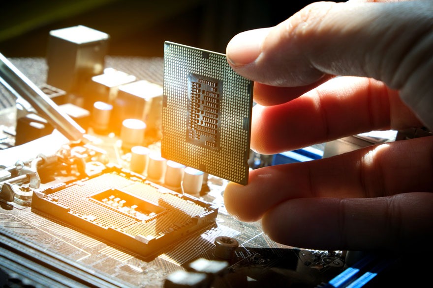 Une main tient un CPU doré au-dessus d'un socket de carte mère. Des composants électroniques sont visibles en arrière-plan.