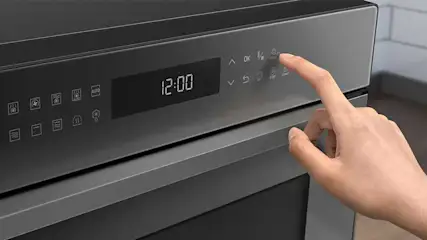 Een grijze oven met een digitaal scherm dat 12:00 weergeeft, wordt bediend door een hand.
