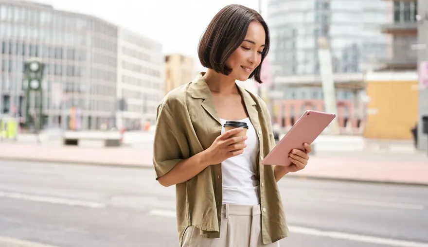 Une personne tient une tablette et un café sur une rue urbaine avec des immeubles en arrière-plan.