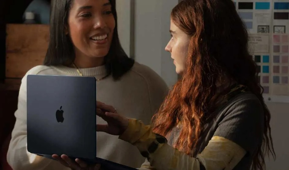Two people chat while viewing a sleek dark‑blue Apple MacBook, its logo visible, in a softly lit studio with colour swatches on the wall.