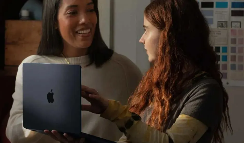 Two people chat while viewing a sleek dark‑blue Apple MacBook, its logo visible, in a softly lit studio with colour swatches on the wall.