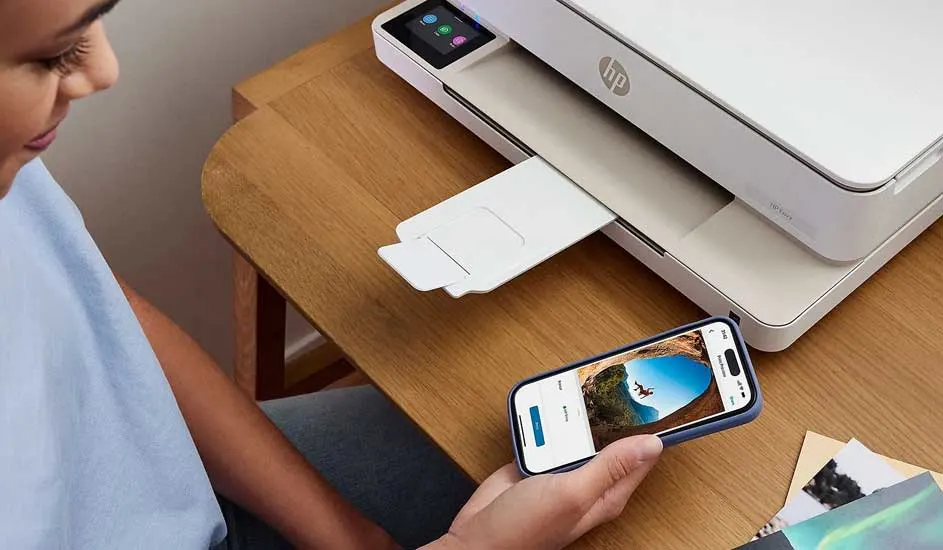 Een witte HP printer staat op een houten bureau. Een smartphone toont een foto. Er liggen foto's op het bureau.
