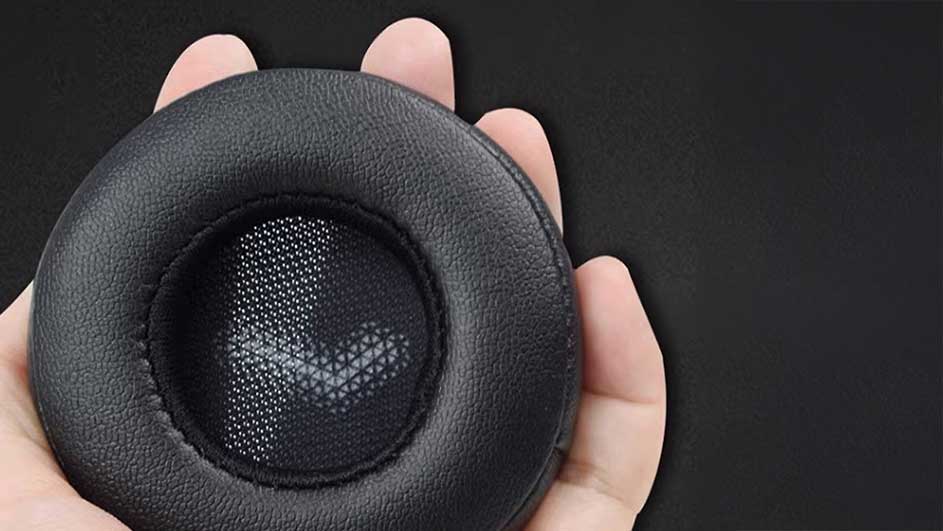 Un coussin d'écouteur noir rond avec un centre en maille grise est tenu dans une main sur un fond noir uni.