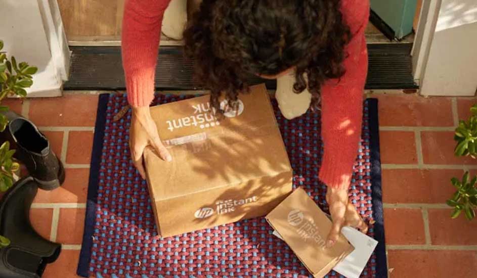 Bovenaanzicht van een deurmat met een bruine doos en envelop erop, schoenen links en een bakstenen vloer.