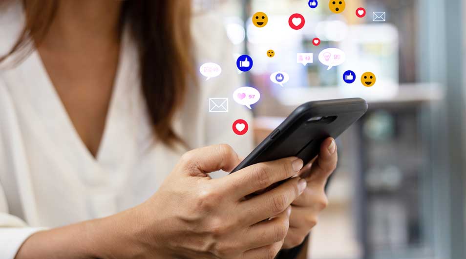Gros plan sur un téléphone noir tenu à deux mains, avec des icônes de réseaux sociaux flottantes. Arrière-plan flou.