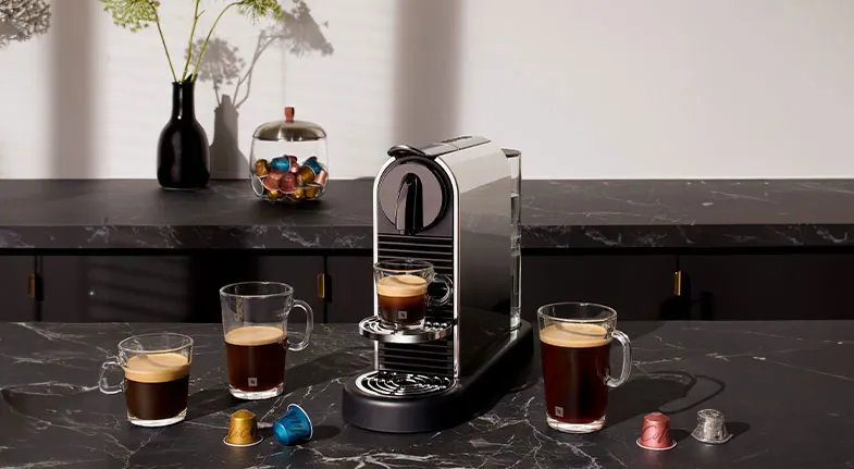 Een zilveren koffiemachine op een zwart marmeren aanrecht, omringd door glazen mokken koffie en capsules.