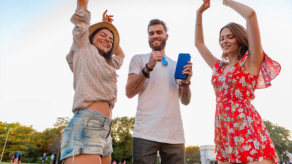 Drie mensen dansen op gras met een blauwe speaker. Bomen en meer mensen zijn te zien op de achtergrond.