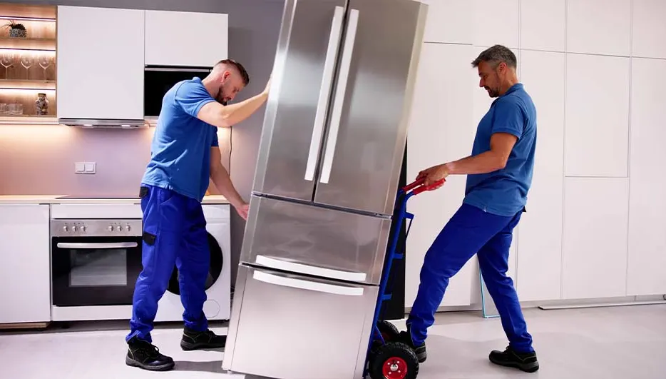 Deux manutentionnaires en uniforme bleu déplacent un réfrigérateur en acier inoxydable dans une cuisine blanche.