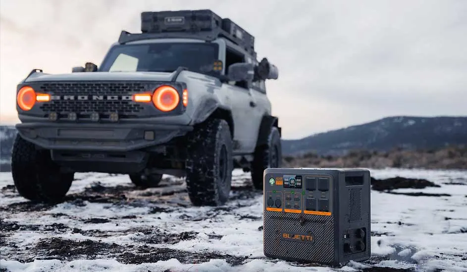 Une station d'énergie BLUETTI est posée dans la neige près d'un 4x4 gris. La BLUETTI a des touches orange et plusieurs ports.