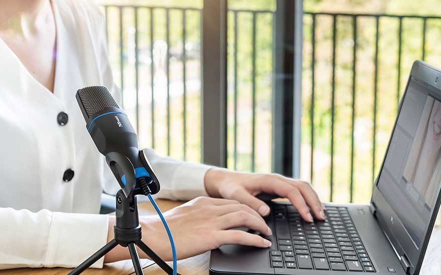 Een zwarte microfoon op een statief staat naast een laptop. Beide staan op een houten tafel. Achtergrond: een raam met verticale lamellen.