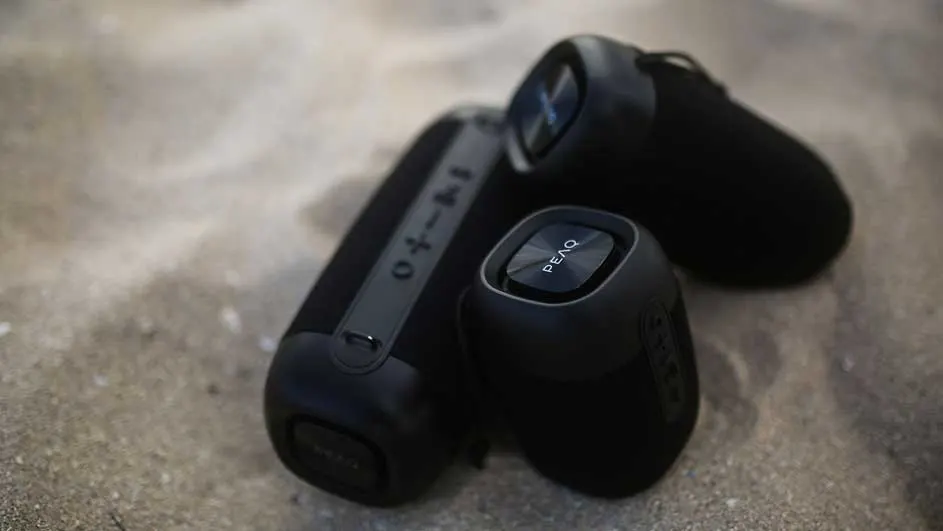 Drie zwarte PEAQ Bluetooth-luidsprekers liggen op een zanderige ondergrond. Verschillende formaten, rechthoekig en rond.