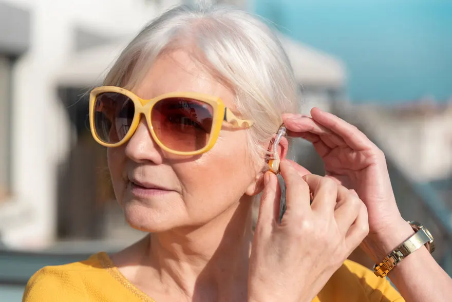 Une personne ajuste un appareil auditif transparent. Elle porte des lunettes de soleil jaunes et une chemise jaune.