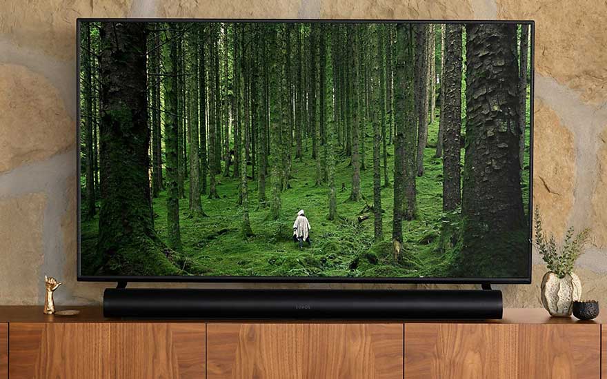 Een tv toont een dicht bos met een persoon in een witte poncho. De tv staat op een houten kast met een zwarte soundbar.