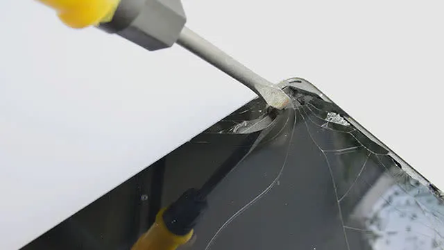 Een geel handvat van een gereedschap steekt in een gebarsten zwart scherm, tegen een witte achtergrond.