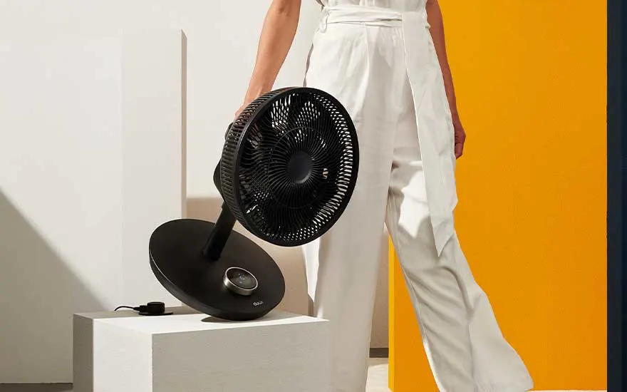 Een vrouw in witte kleding houdt een moderne zwarte ventilator vast, geplaatst op een witte sokkel voor een gele muur.