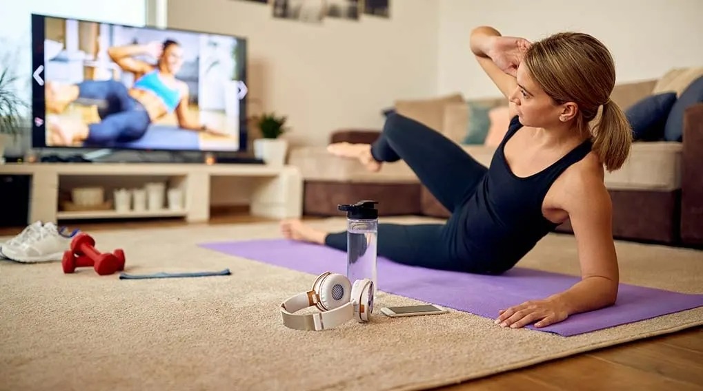 A person does abs workout on a purple mat in front of a TV. Nearby are headphones, a water bottle, red dumbbells, and a phone.