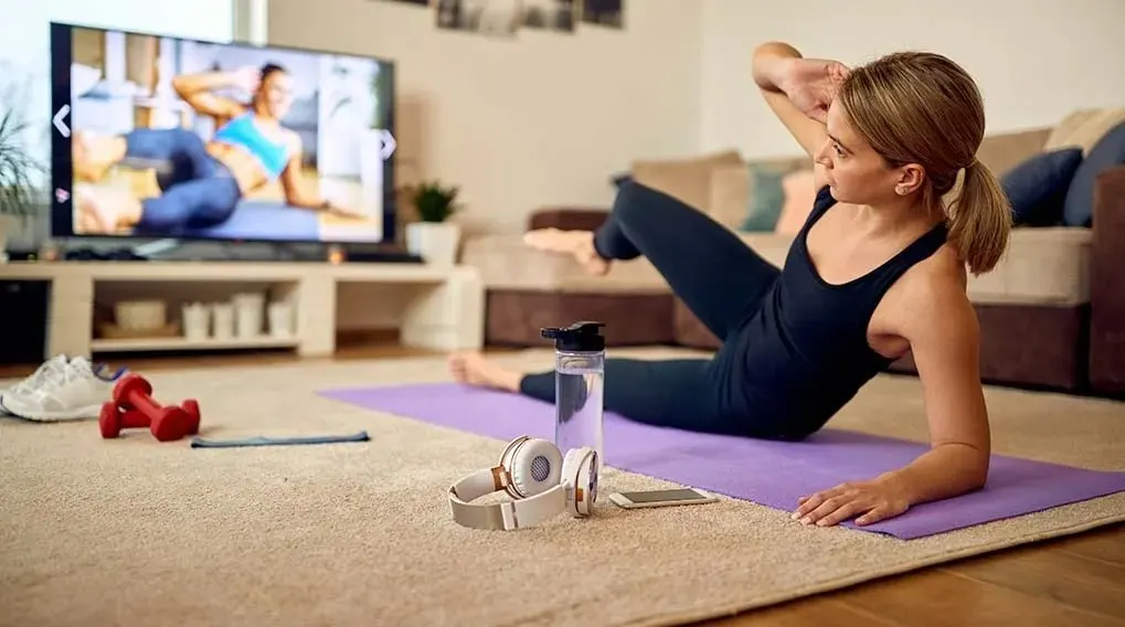 A person does abs workout on a purple mat in front of a TV. Nearby are headphones, a water bottle, red dumbbells, and a phone.