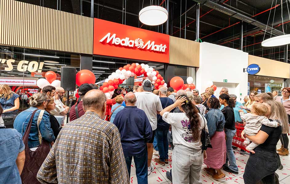 In een drukke MediaMarkt-winkel staan veel klanten rond een boog van rode en witte ballonnen. Sommigen dragen kinderen, terwijl personeel helpt.