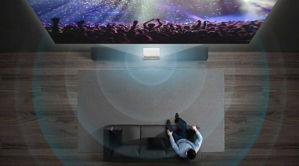 Een woonkamer met houten vloer, een grijze zetel en een projector die een concert projecteert op de muur.
