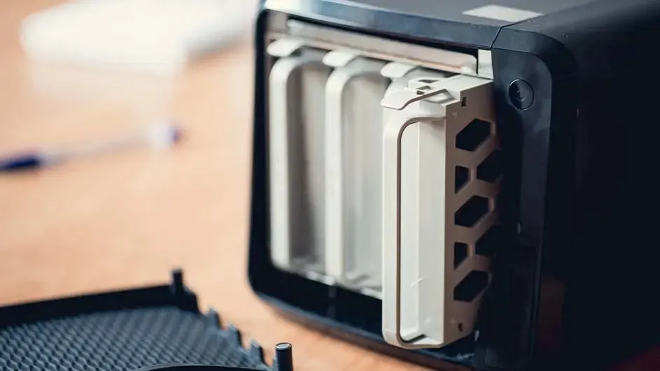 Boîtier noir avec baies internes blanches visibles et un support latéral blanc perforé. Sur une table en bois.