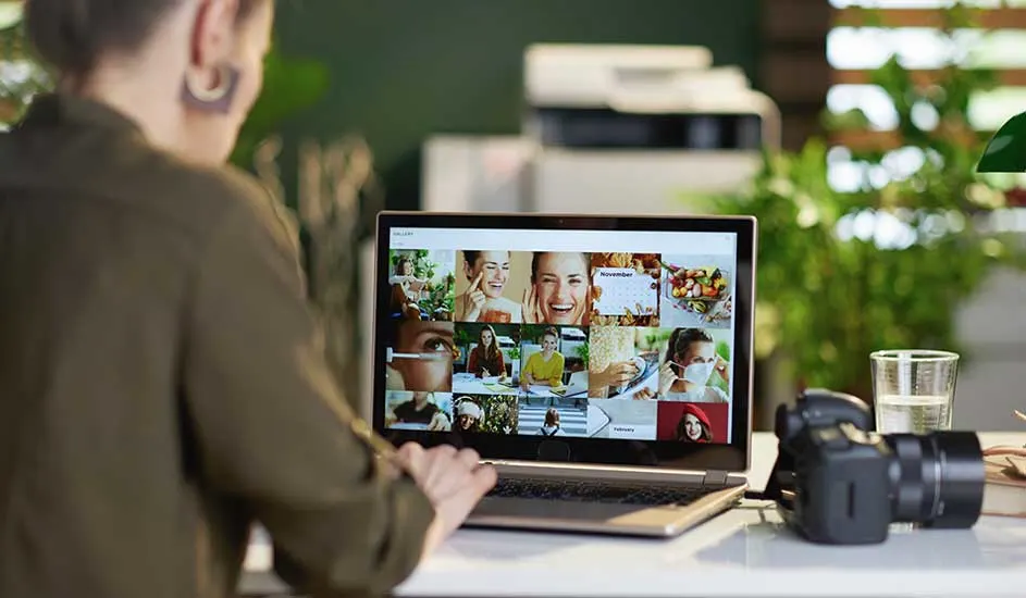 Person reviews a colorful photo gallery on a laptop, with camera and glass of water on the desk amid lush green plants.
