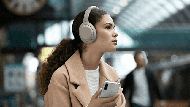 Une femme porte un casque beige et tient un smartphone blanc. En arrière-plan, une gare est visible.