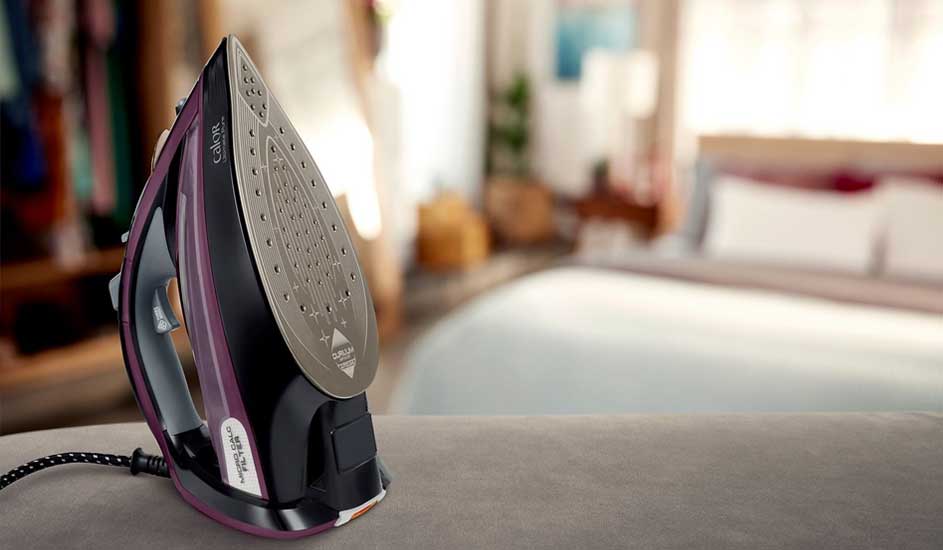 Un fer à repasser noir et violet sur une surface grise, avec un lit et une armoire en arrière-plan flous.