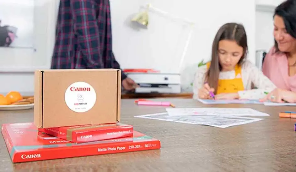 Een bruine doos met een Canon-logo staat op rood Canon-fotopapier. Op de achtergrond staat een printer op een tafel.