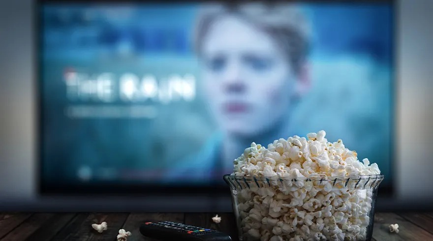 A bowl of popcorn and a remote sit on a table in front of a TV screen showing "The Rain".