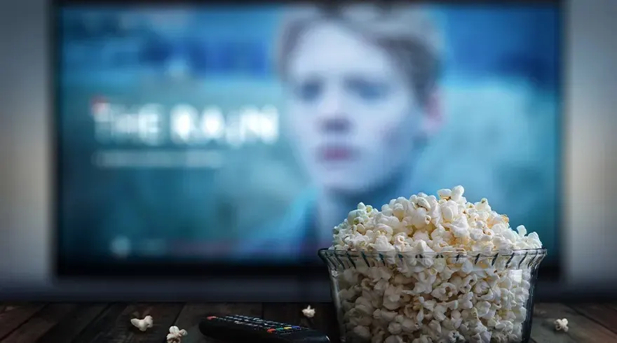 Un bol de pop-corn et une télécommande devant un écran de télévision allumé sur une table en bois sombre.