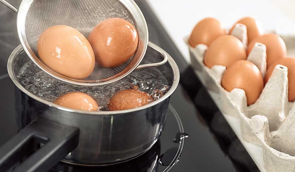 Eieren in een kookpot met kokend water, een zeef erboven. Eierdoos rechts op een zwart oppervlak.