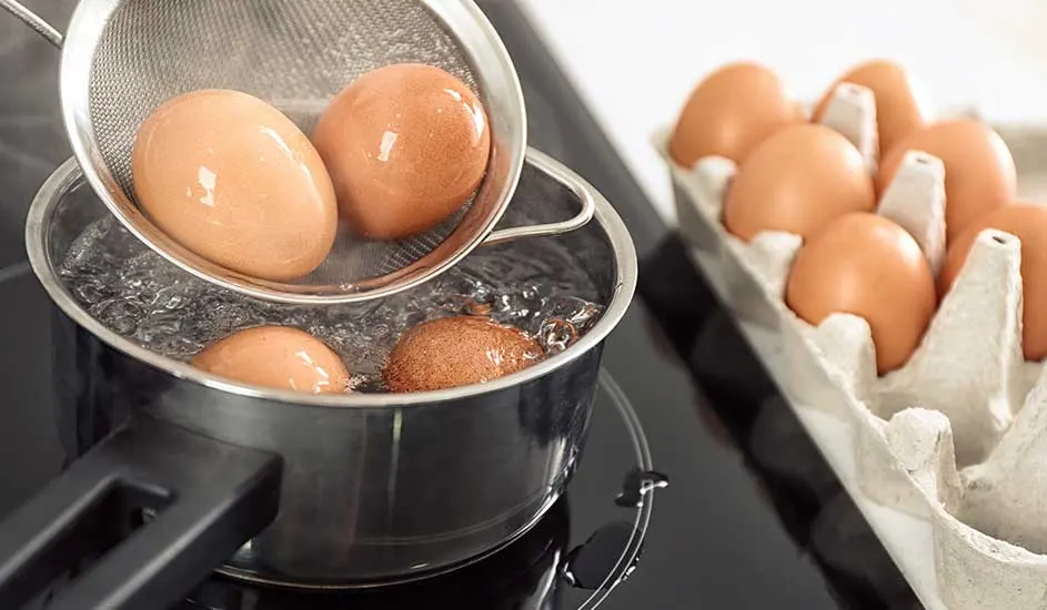 Des œufs bruns dans une casserole d'eau bouillante et dans une boîte à œufs en carton sur une plaque de cuisson noire.