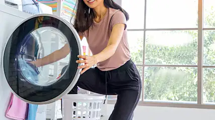 Une machine à laver blanche avec un hublot rond à bord noir. Un panier à linge blanc devant une fenêtre.
