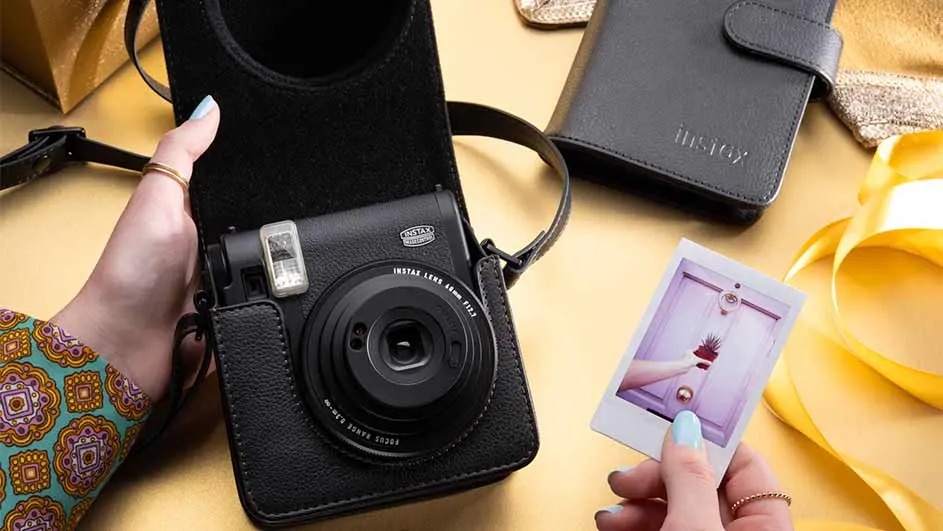 Une personne tient un appareil photo noir dans son étui et une photo instantanée sur une table avec des cadeaux et un ruban.