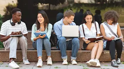 Vijf studenten zitten samen buiten, maken aantekeningen en gebruiken een laptop om te studeren.