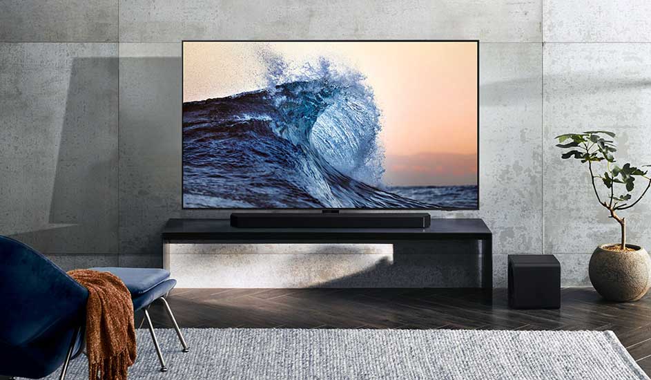 Un téléviseur Samsung affiche une vague à l’écran. Une barre de son et un caisson de basses Samsung sont posés sur un meuble noir dans un salon moderne et lumineux.