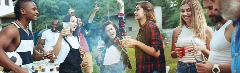 A group gathers outdoors, holding drinks. A soccer ball is visible. Trees are in the background.