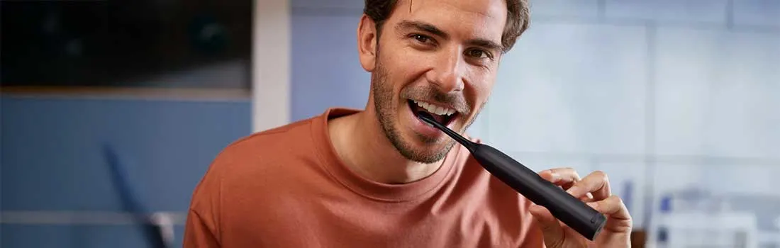 Een man in een rood shirt poetst zijn tanden met een zwarte elektrische tandenborstel in een modern ingerichte badkamer.