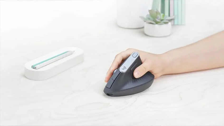 Une main manipule une souris verticale noire. Un stylo blanc repose à côté. À l'arrière, une petite plante et des vases.