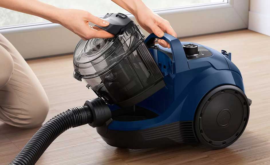 Un aspirateur bleu sur un plancher en bois, avec une personne retirant le réservoir à poussière transparent. Fenêtre en arrière-plan.