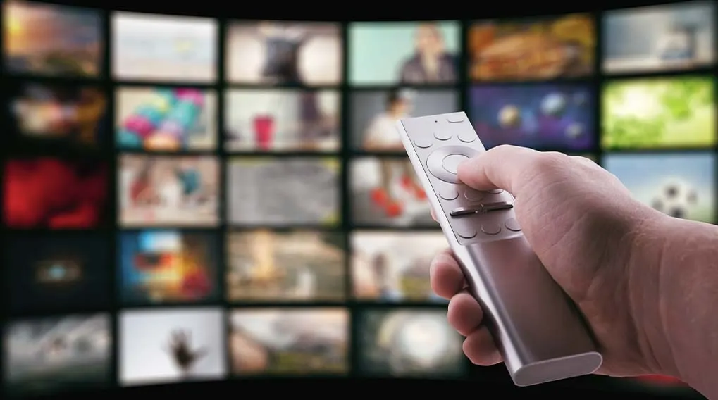 Une main tient une télécommande argentée. Un mur d'écrans de télévision flous est visible en arrière-plan.