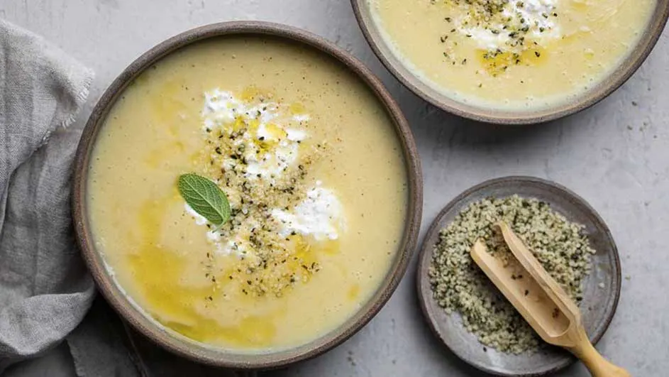 Deux bols de soupe jaune crémeuse sont garnis de crème, d'herbes et d'une feuille verte. Une assiette de graines et une cuillère en bois sont à côté.